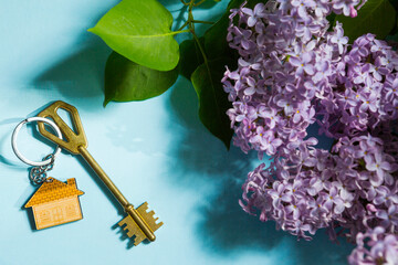House key with keychain on blue spring background and lilac branches.Summer holiday home, booking cottage in the countryside, moving to new home, mortgage, rent and purchase real estate. Tourism