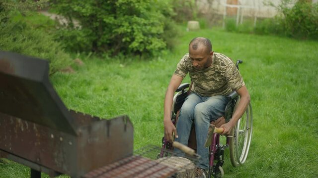 Disabled military veteran chopping wood in slow motion on backyard at home. Wide shot of serious handsome African American man in wheelchair preparing picnic outdoors