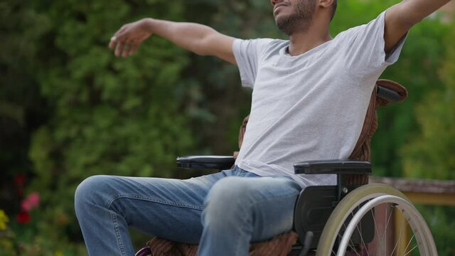 Unrecognizable disabled man in wheelchair stretching hands sitting outdoors on spring summer day. Optimistic African American paralyzed invalid enjoying leisure. Confidence and lifestyle
