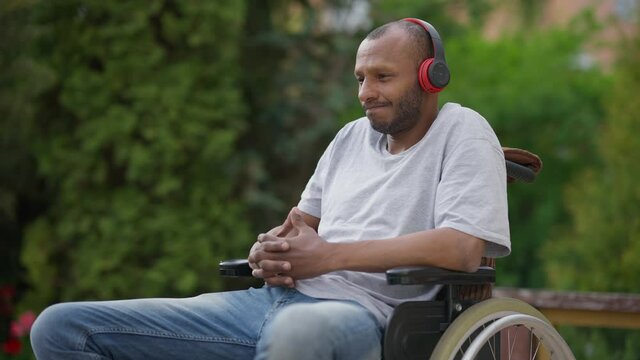 Medium shot of positive African American paraplegic man in wheelchair enjoying music in headphones. Portrait of relaxed thoughtful disabled person listening to playlist in earphones outdoors