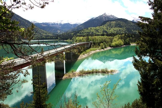 view down over highway bridge with mountains and green forest behind and green turquoise lake water with sky reflexions of mountains and clouds on surface © Andreas