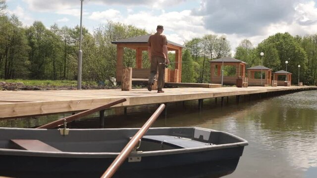 Man walking on wooden pantoon near lake with boat in summer. Fisherman going along water with fishing equipment.