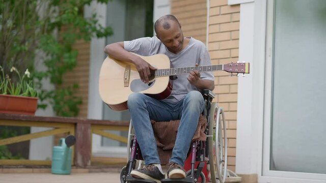 Portrait Of Disabled African American Musician Guitarist In Wheelchair Sitting On Porch With Guitar Touching Strings. Front View Wide Shot Of Positive Confident Crippled Man Enjoying Hobby Outdoors