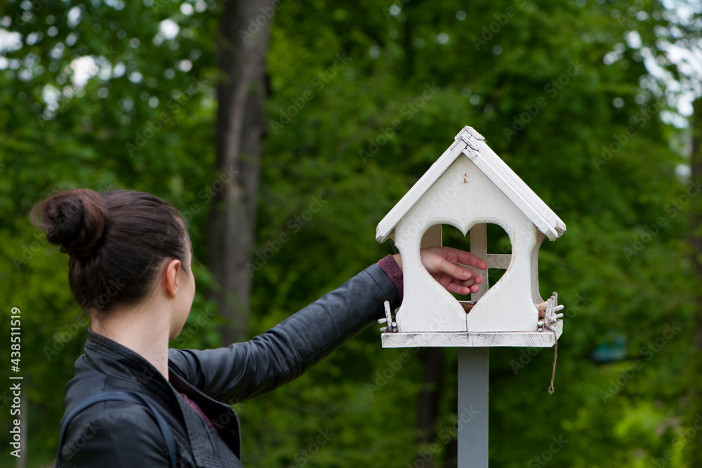 Wall mural feeds birds in a feeder in an spring park. girl puts food. girl out of focus, rear view. taking care of birds, natural green blurred background. wooden birdhouse. white feeder in the garden. close-up