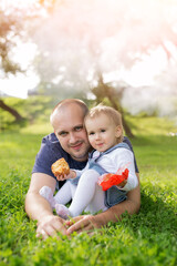 Fototapeta premium A little girl is eating a pie in her father's arms and laughing merrily. The relationship between father and daughter. Picnic in the park at sunset. A loving father. Happy family. Childhood in harmony
