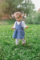 A little girl walks on the grass with her hands raised and a sly look on her face. The child takes the first confident steps