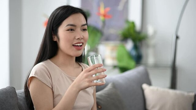 Healthy pretty asian brunette girl, holding and drinking a glass of pure water, while sitting at home on sofa in casual t-shirt, leading a healthy lifestyle, smiling