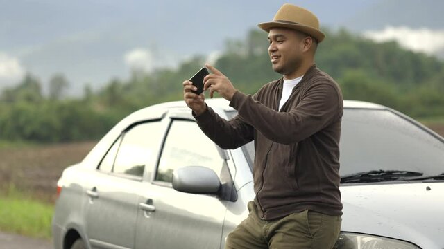 A young Asian man drives car to beautiful nature. He was standing in front of the car on the roadside. He uses his phone to take pictures of beautiful views.