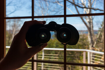 Hand holding binoculars looking out window toward lake.