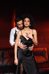 Man hugging african american woman in necklace and dress on black background with red lighting