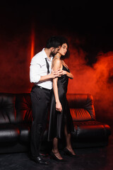 Man in formal wear kissing neck of african american girlfriend on black background with red lighting and smoke