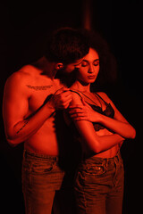 Muscular man taking off bra from sensual african american woman in red light on black background