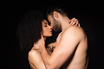 Side view of shirtless man kissing african american woman isolated on black