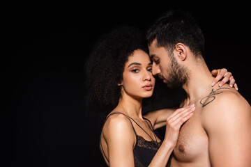 Sexy african american woman looking at camera while embracing shirtless man isolated on black