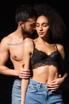 Shirtless and bearded man embracing african american woman in bra isolated on black