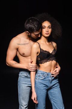 Shirtless man kissing african american woman in jeans and bra isolated on black