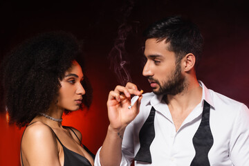Man smoking cigarette near elegant african american girlfriend on black background with red lighting