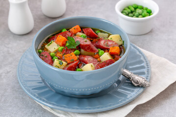 Smoked sausages, carrot and potato stew served in blue ceramic bowl with fresh garden pea and parsley. Light concrete background with copy space, selective focus.