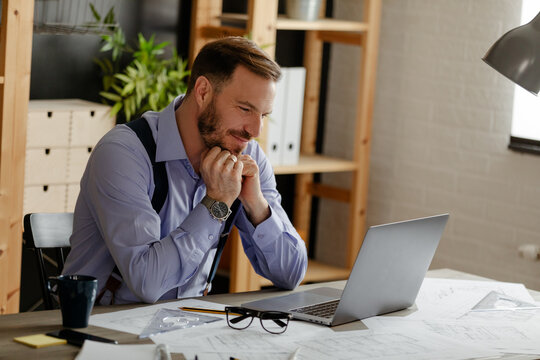 Young Smiling Man Working At Home On His Laptop Typing, While He Is Surrounded By Papers