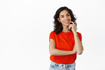 Stylish brunette girl with curly hairstyle, red t-shirt, biting finger coquettish while thinking, making choice or decision, standing over white background