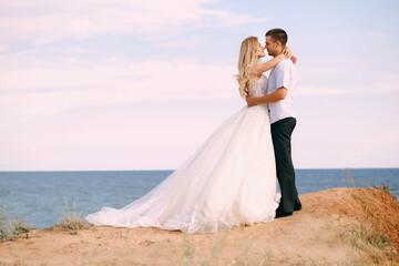 Fototapeta na wymiar The bride and groom hug on the edge of the mountain against the background of the sea. An elegant bride in a white wedding dress looks tenderly at her husband. Happy newlyweds hug. Free space for text