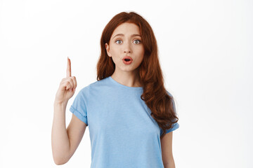 Excited redhead female model in t-shirt, pointing finger up and say wow, showing interesting promo offer, demonstrating advertisement on top, standing against white background