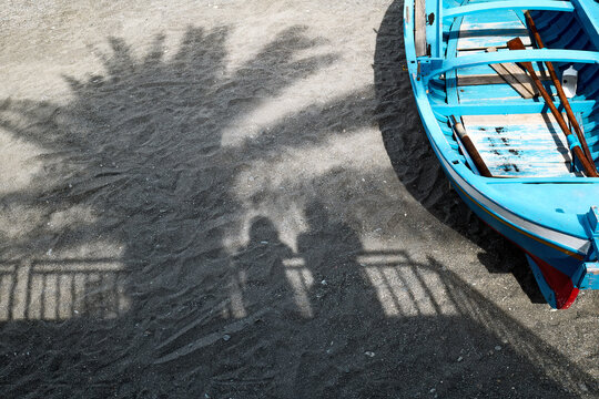 Shadow of two tourists under a palm tree on a pabble beach. Blue boat near silhouette of a couple. Summer time vacation on the sea, tourism concept.