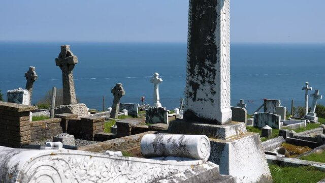 Slow tilt up shot of Celtic Christian ringed cross in Welsh cemetery on seaside hill with blue sea and sky background.