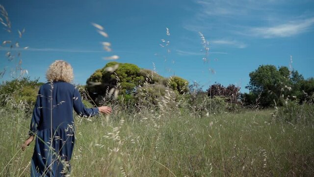 A European woman, middle-aged in a dress, walks on a prak with tall grass. With a philosophical romantic mood. Slow motion