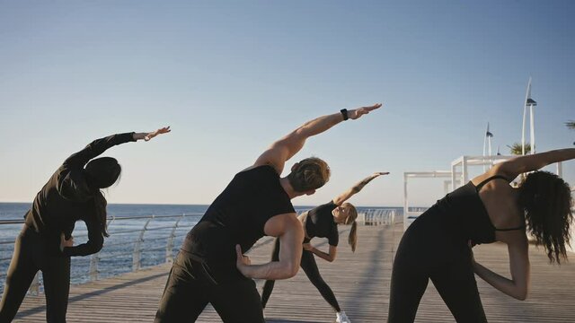 Young woman fitness coach showing stretching exercise to group of sporty people, practicing training on pier, zoom out