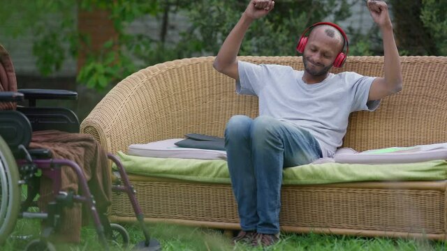 Wide Shot Of Relaxed Carefree Handicapped Man Enjoying Music In Headphones Moving In Slow Motion Sitting On Couch In Garden. Portrait Of Positive Smiling Happy African American Disabled Guy Outdoors