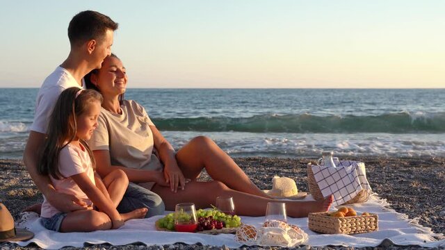 Happy family have a picnic on the beach at sunset. Mom, dad and daughter are resting seaside at summer vacation.