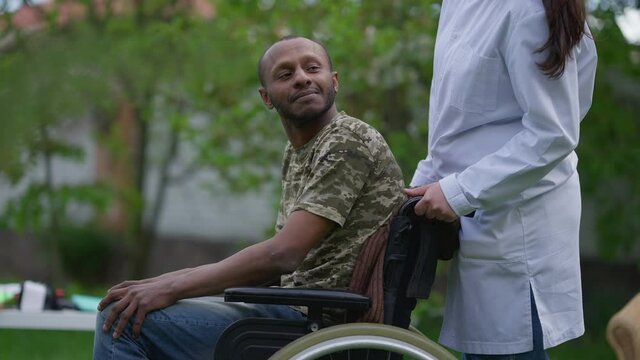 African American Paralyzed Man Greeting Unrecognizable Caucasian Nurse Pushing Wheelchair Leaving. Young Caregiver Taking Care Of Disabled Patient In Nursing Home Garden Outdoors