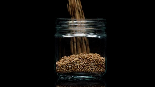 Close-up of falling down buckwheat into glass jar on black background. Shooting of cereal and groats in studio. Macro footage of pouring out the grits. Food cooking video. 