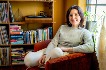 Portrait of smiling woman sitting in chair next to bookshelf and window 