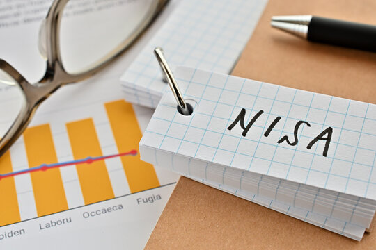 There is a word book with the word of NISA which is an abbreviation for Nippon Individual Savings Account on the desk with papers of graphs(with dummy text), a pen and glasses.