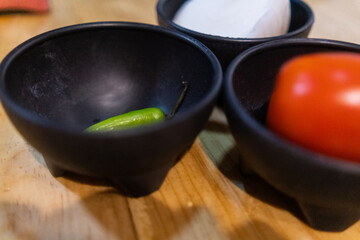 Tomatoes, onions, and chili peppers in black bowls above wooden surface