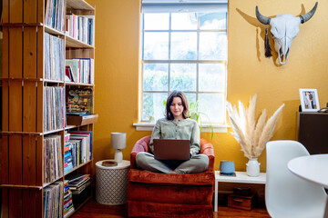 Woman sitting in orange chair by bookshelf and window working on laptop