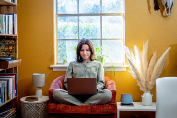 Woman sitting in orange chair by bookshelf and window working on laptop