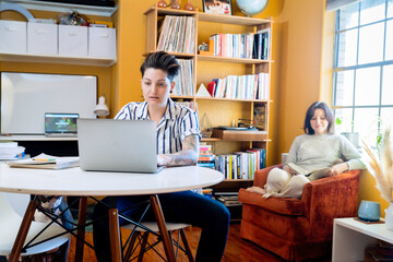 Woman working on laptop at table while wife reads in background