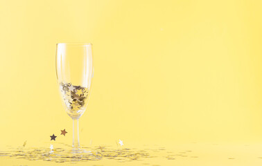 A glass of champagne filled with gray stars on a yellow background. Gray stars hover in the air around the case. New Year Party and celebration concept with large copy space