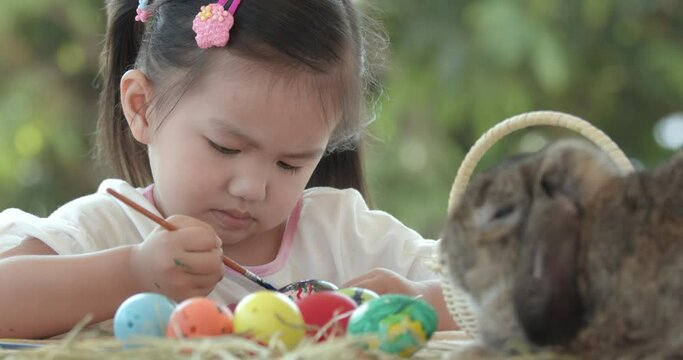 Close up of asian little girl coloring easter eggs with colors and brush near a brown rabbit on easter festive desk. Preparation of the Easter. Zoom in, slow motion