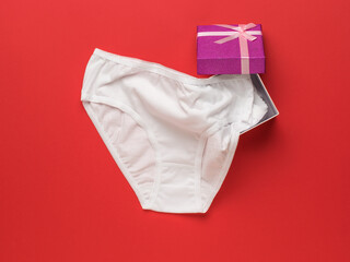 White women's panties sticking out of a gift box on a red background. Flat lay.