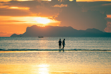 Fototapeta premium Young couple walks on a sand in the sea against the backdrop of islands and a colorful sunset and enjoys a summer evening 