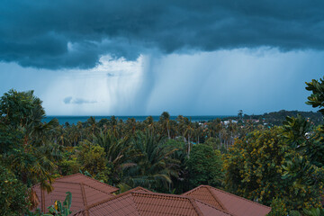 Rain and storm comes over a forest valley, sea and rooftops on a summer day on a tropical island. View from the balcony