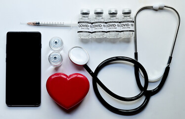 Top Views of Coronavirus COVID-19 Vaccine Glass Bottle, syringe, Black stethoscope, Heart-shaped plastic box and Modern Black Smartphone isolated on blue background. Medical device concept.
