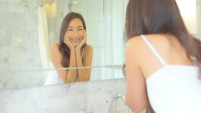 Beautiful Smiling Asian Woman Inspects her Perfect Face in the Mirror.