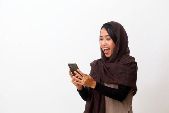 Portrait of surprised asian muslim woman wearing a veil or hijab using a cell phone and smiling. Receiving good news. Isolated on white background with copy space