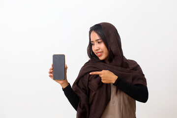 Portrait of happy asian muslim woman wearing a brown veil or hijab and white dress showing and pointing a blank smartphone screen. White background with copy space