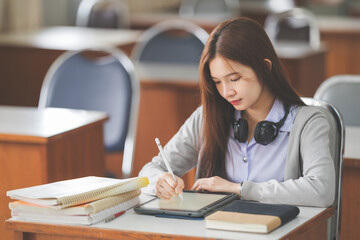 Stock photo of a young teenage woman Asian college student in student uniform studying and writing on digital tablet in a university classroom